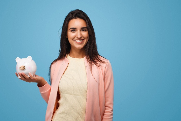 Brunette woman wearing a pink cardigan holds up a white piggy bank for saving money against a blue wall