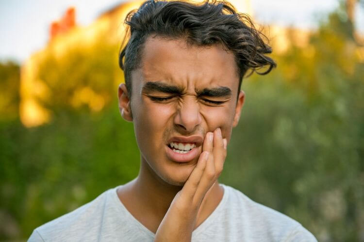 Dark haired teenage boy is outdoors with forehead wrinkled, eyes closed and hand to jaw experiencing tooth pain