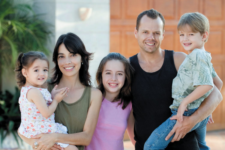 Family in front of a house with the mom holding a toddler girl, dad holding a young boy and a young adolescent girl standing between them