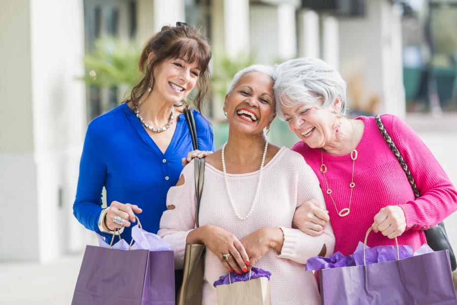 Three smiling mature women walking down the street together arm in arm with shopping bags.